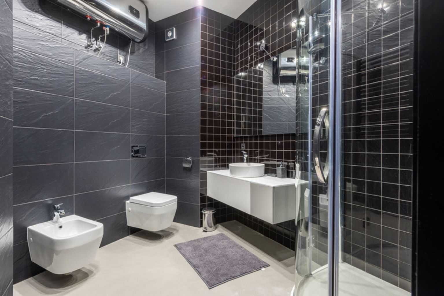 Badeværelse med mørke vægfliser, væghængt toilet og bruseniche med glasvæg