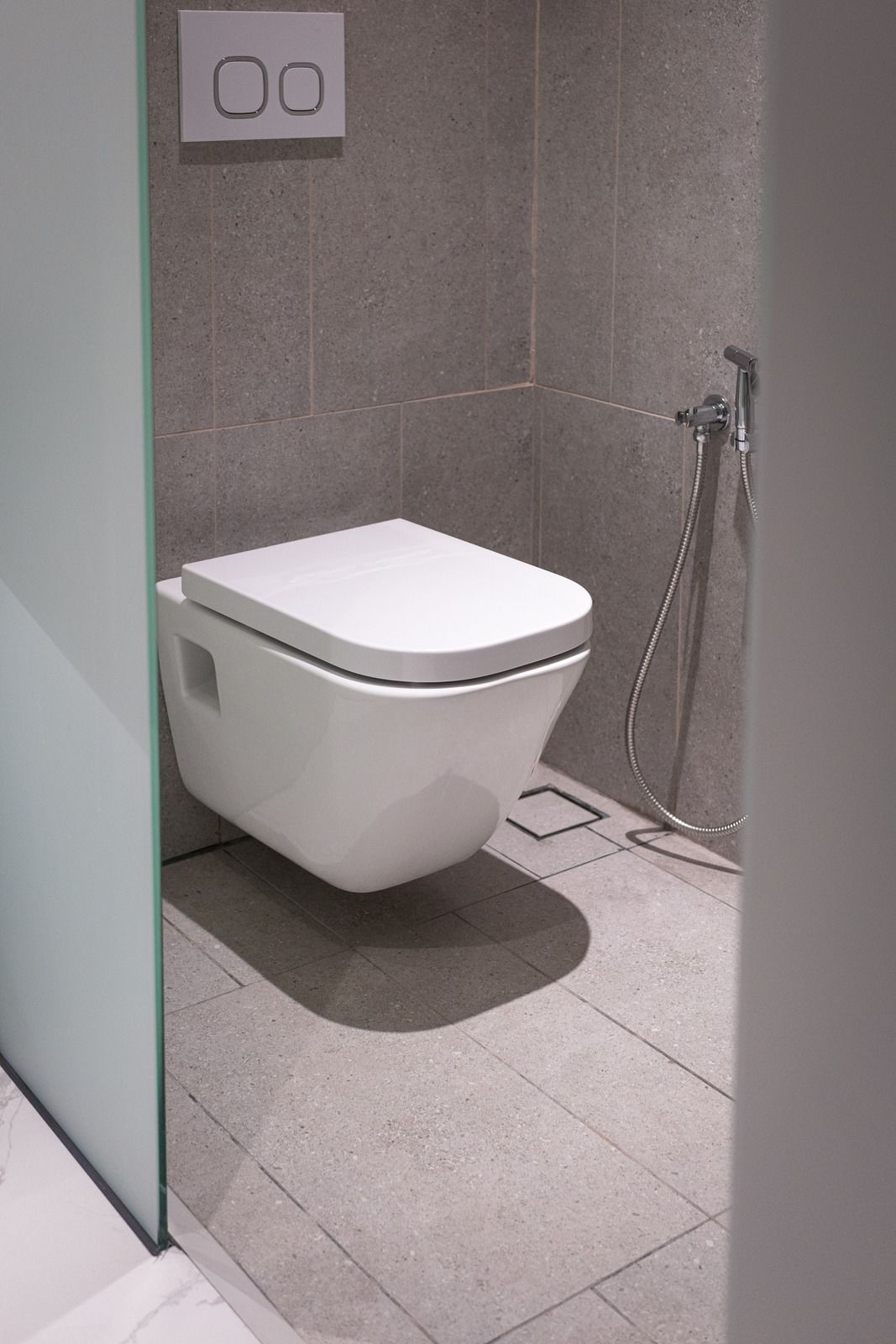 Hvidt firkantet væghængt toilet med betjeningsplade på grå væg og håndbruser ved siden af