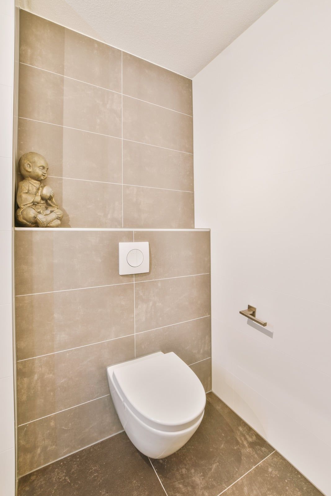 Hvidt væghængt toilet med betjeningsplade på væg med grå fliser og en hylde over indbygningen